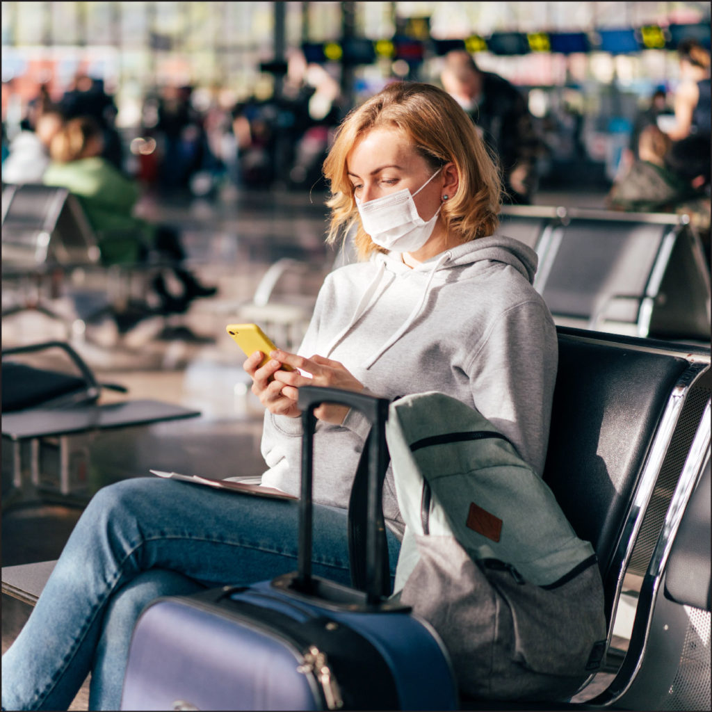 Image of a masked woman sitting at an airport looking at her phone, with luggage in front of her