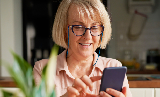 A smiling woman with short blond hair and glasses is looking at her smartphone. She is indoors, with a blurred background that includes a plant and assorted household items. She appears to be using her right hand for appointment scheduling on an AI-enabled outreach app.